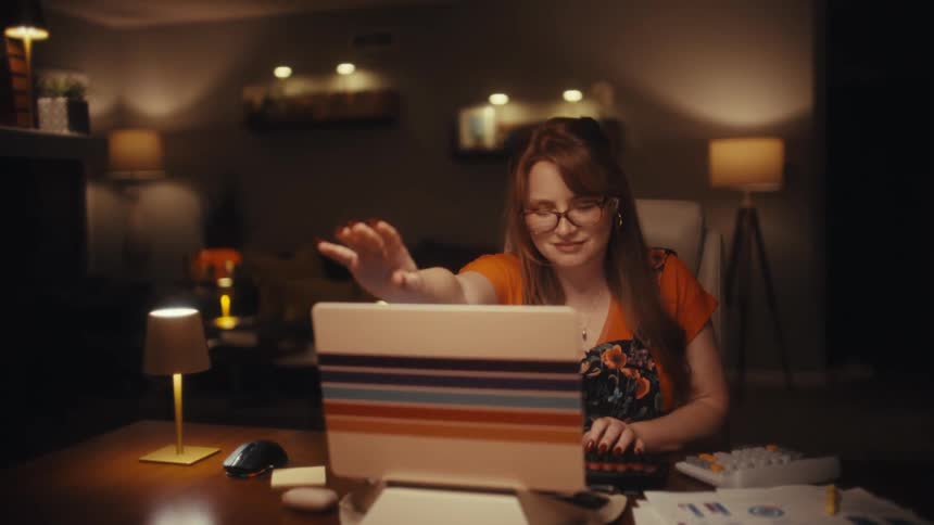 In a warmly lit home office at night a focused woman wearing glasses works at a laptop before closing the laptop in a low light ambient room