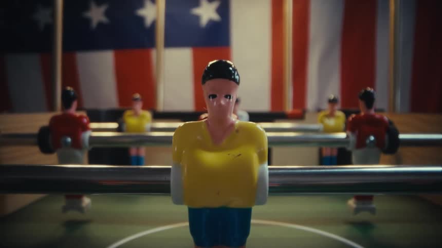 Table football game closeup showing plastic players in yellow blue and red uniforms positioned in front of a large american flag backdrop