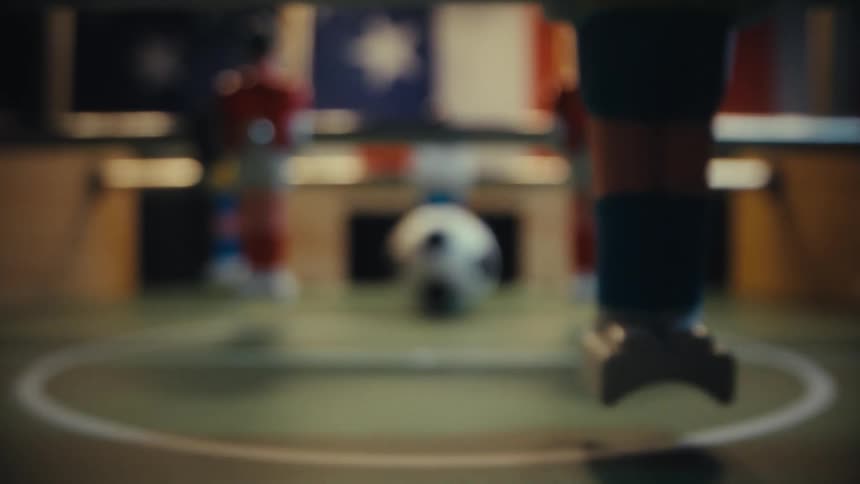 Starting with a sharp closeup on a stationary black and white ball the camera pulls backward across the green surface of a foosball table passing the plastic legs of a game figure