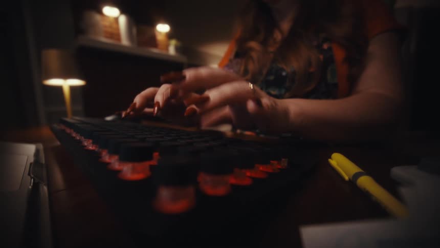 A closeup shot focuses on a womans hands with long wellmanicured dark fingernails as she types rapidly on a mechanical keyboard featuring round glowing orange keys in a dimly lit setting