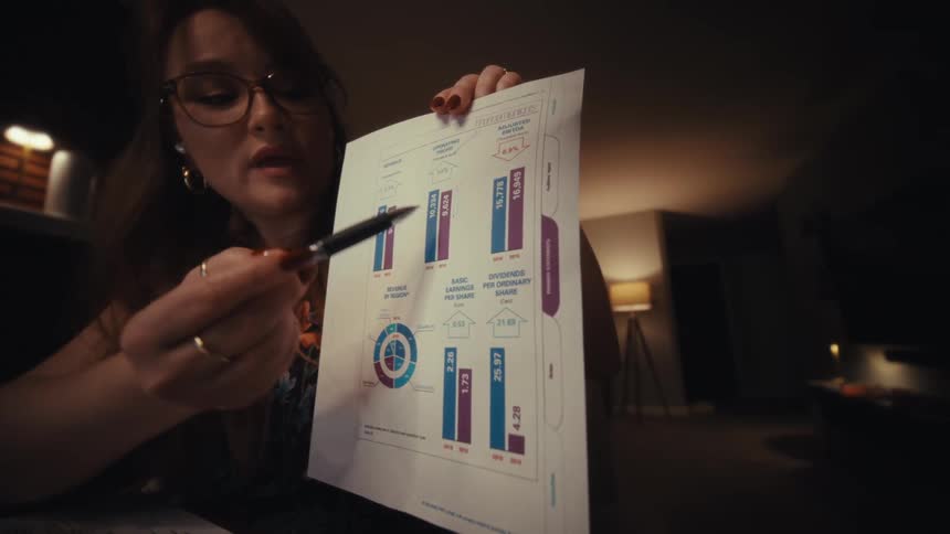 A woman wearing glasses holds up a detailed chart and points to a specific section with a pen her focused expression and the warm softly lit room emphasizing careful analysis and deliberate explanation