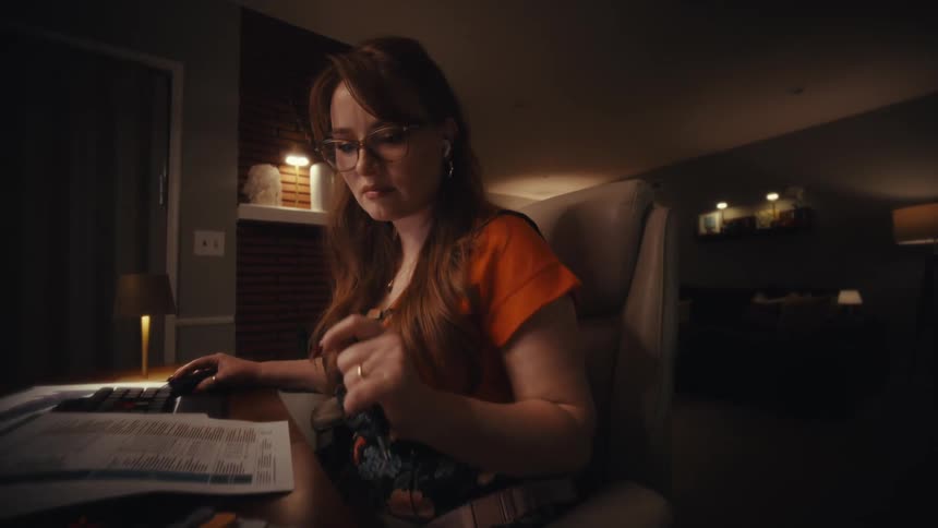 Young adult woman in a living room pressing buttons on a calculator machine over a table covered in sheets of paper indoors during night time