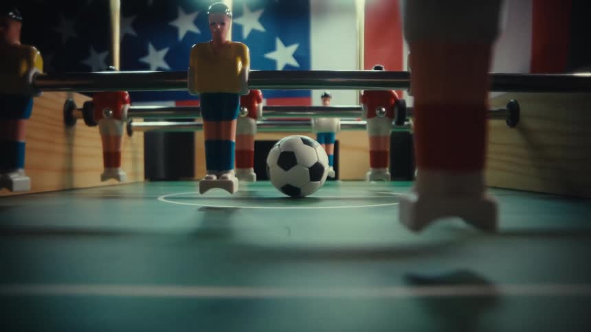 The camera pulls back through a foosball table starting with a closeup of a soccer ball and moving past rows of miniature players in the background an american flag is visible behind the game table as the perspective shifts through the goal opening