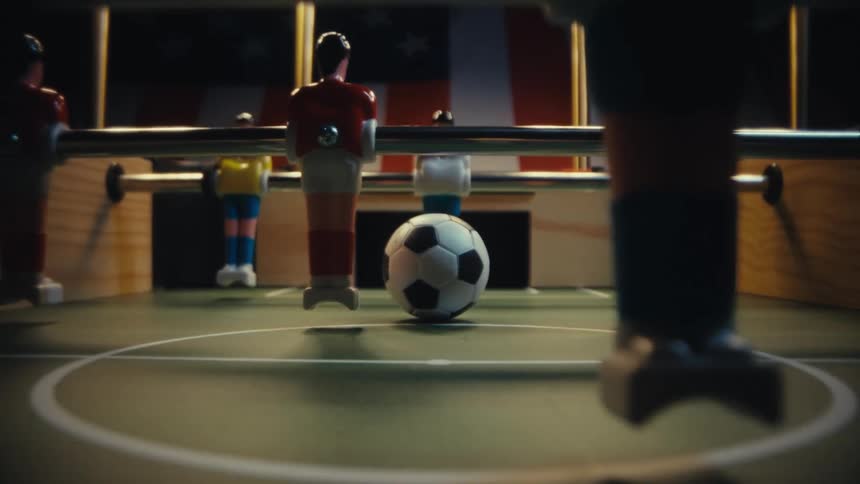 A dramatic lowangle shot follows a foosball at midfield as the camera slowly pans out revealing rigid player figures in colorful uniforms looming around it with polished metal rods
