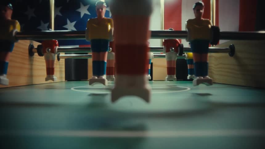 The camera glides through the rows of plastic players on a foosball table eventually stopping on a closeup of the ball at center pitch an american flag hangs in the background behind the table
