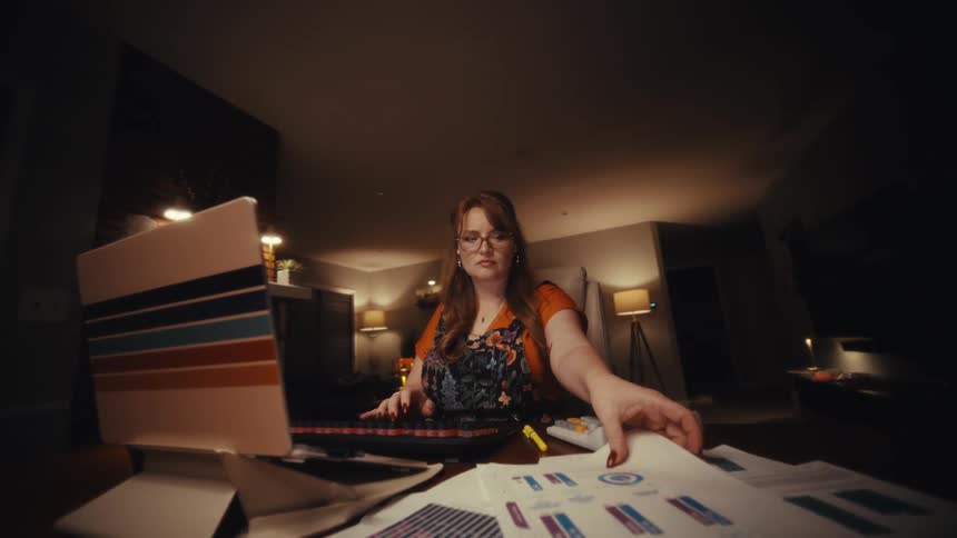In a softly lit home office at night a woman reviews printed documents at a cluttered desk her focused expression and warm lighting conveying concentration and latehour work
