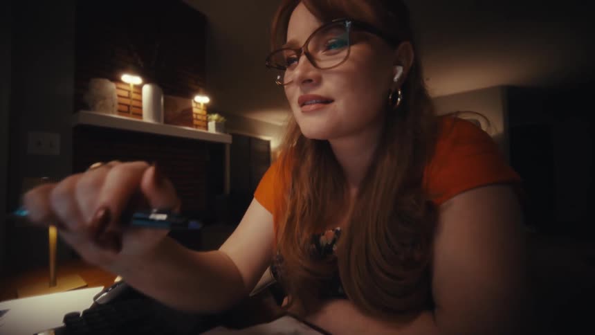 Young woman with red hair and glasses working late at night reviewing business documents and financial charts at her desk in a warmly lit home office