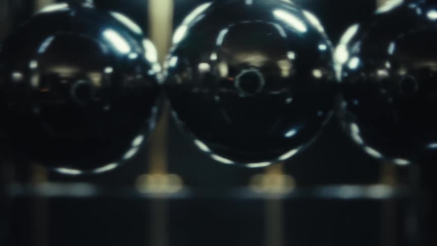 A sleek newtons cradle sits under bright focused lights its polished metal spheres suspended in perfect alignment as reflections ripple across their mirrored surfaces against a dark minimalist background
