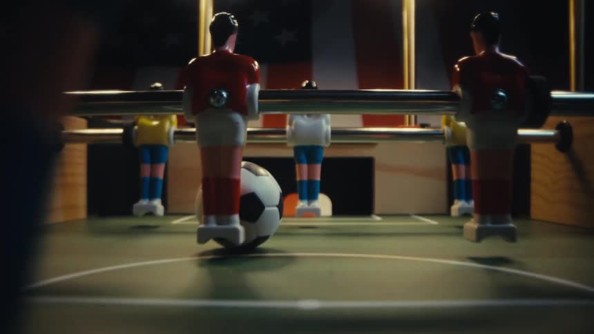A lowangle shot moves through a foosball table passing between plastic players in red and white uniforms toward a miniature soccer ball while an american flag hangs in the dark background behind the wooden goal
