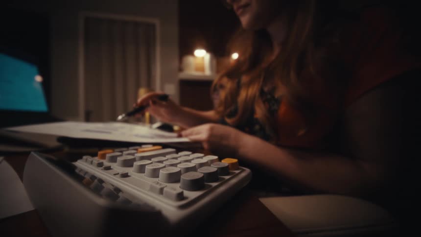 In a dimly lit room a woman works late at her desk using a pen and a calculator with round retro style keys
