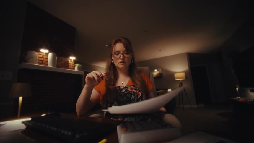 A woman wearing glasses and an orange shirt sits at a desk late at night carefully reviewing printed charts and financial documents the scene shifts from a wide shot of her cluttered workspace to a closeup as she leans in appearing focused and slightl