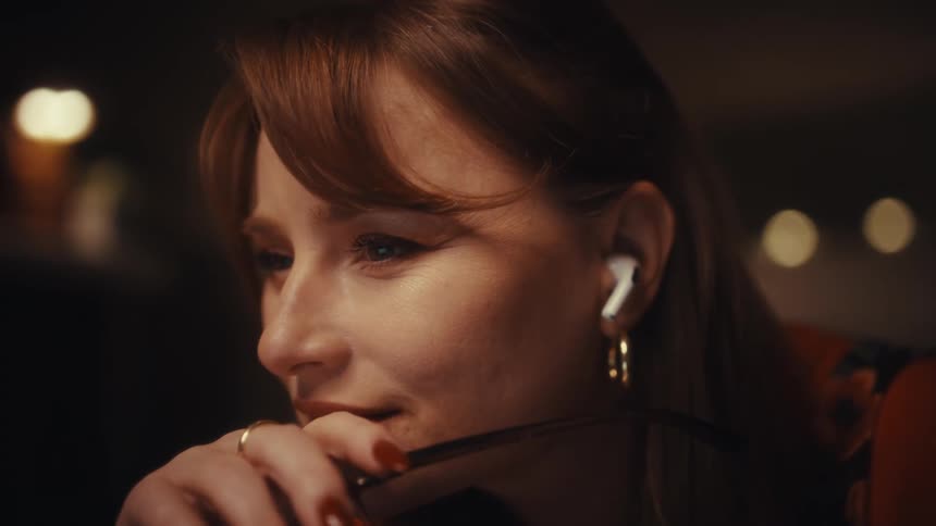 A woman wearing an orange top and wireless earbuds removes her glasses while looking at a screen she smiles thoughtfully and puts her glasses back on as she continues to focus on her work