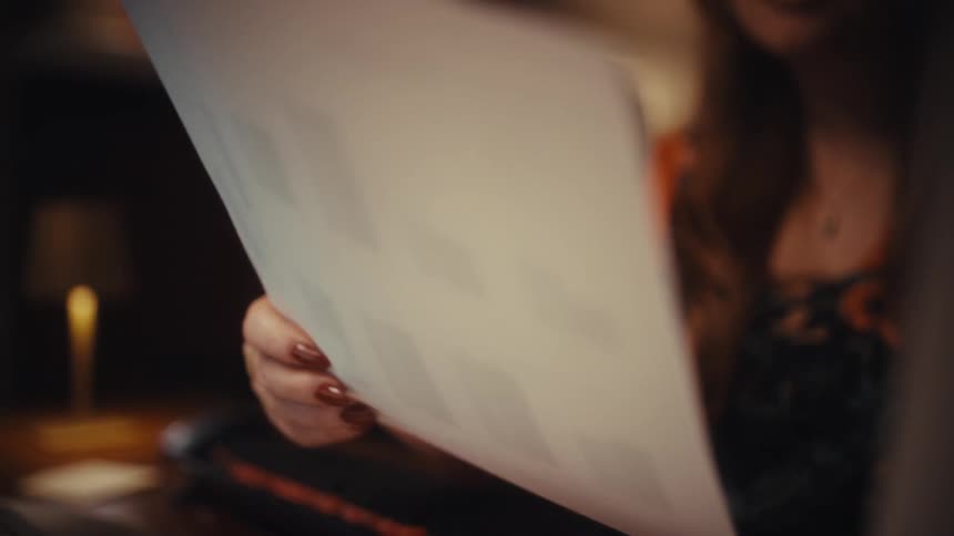 Young adult woman wearing glasses sitting down and reading while holding sheets of paper on her hands indoors during night time