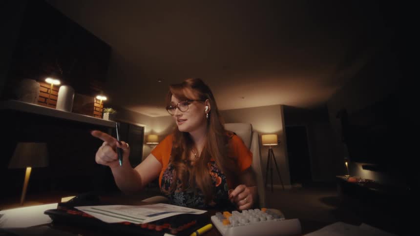 A woman in an orange shirt and glasses sits at a desk at night reacting with excitement and animated gestures while looking at her laptop screen her workspace is covered in financial charts and documents and she appears to be celebrating a breakthrough
