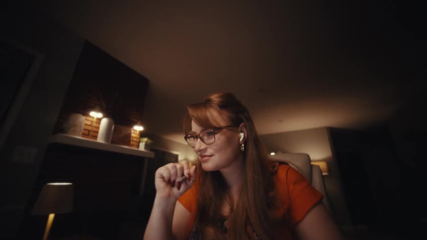 Professional woman with red hair and glasses working in a dimly lit home office smiling while reviewing financial charts and documents on her desk under warm ambient lighting