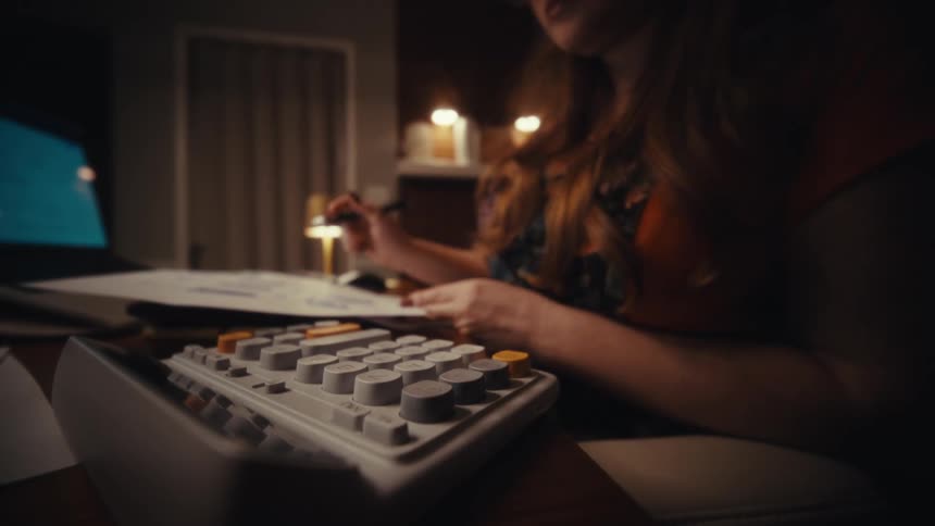 A woman with long red hair wearing an orange shirt and glasses uses a calculator and takes notes while working late at a desk in a dimly lit room