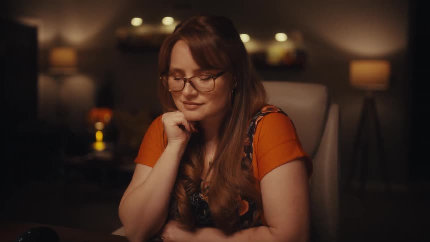 A woman with glasses types on a mechanical keyboard in a dimly lit home office then closes her laptop and rests her chin on her hand with a thoughtful expression while sitting at a desk with charts and a lamp