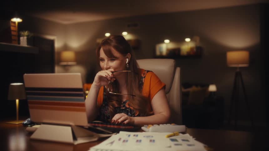 In a dimly lit home office a woman types on her laptop before pausing to remove her glasses and smile thoughtfully at her work scattered charts and warm lighting surround her as she eventually leans back in her chair with an expression of quiet satisfac