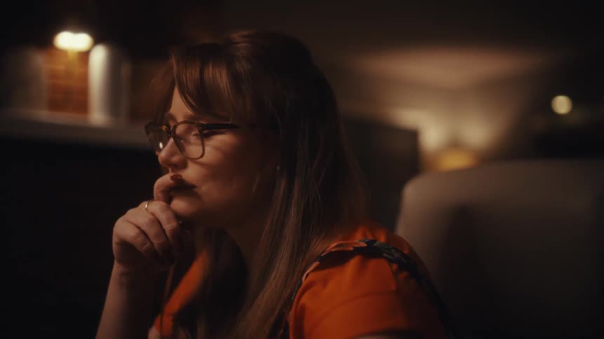 In a dimly lit room a woman sits at her desk and focuses intently on her computer screen while typing she pauses her work to thoughtfully rest her chin on her hand a slight smile appearing on her face