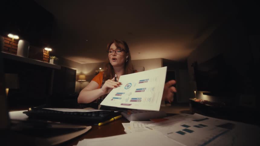 A woman wearing glasses and wireless earbuds sits at a desk cluttered with documents engaging in a professional video call via her laptop she holds up various charts and graphs pointing to specific data points with a pen as she explains them during the