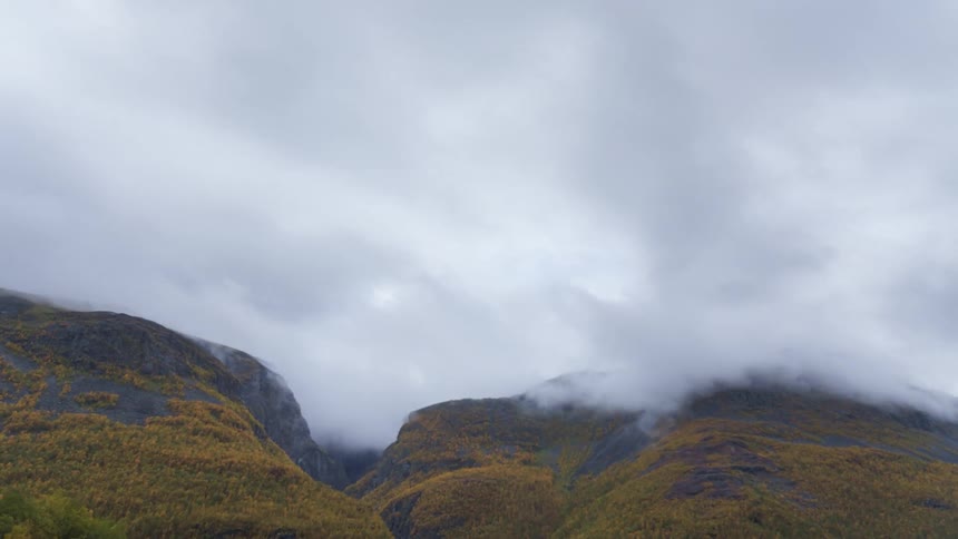 Thick lowhanging clouds drift slowly across the peaks of a vast mountain range covered in yellow autumn trees and dark rock faces under an overcast sky