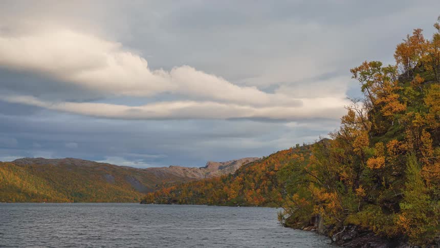 The camera pans right across a large lake surrounded by hills covered in yellow and orange autumn trees under a cloudy sky revealing a steep rocky embankment in the foreground
