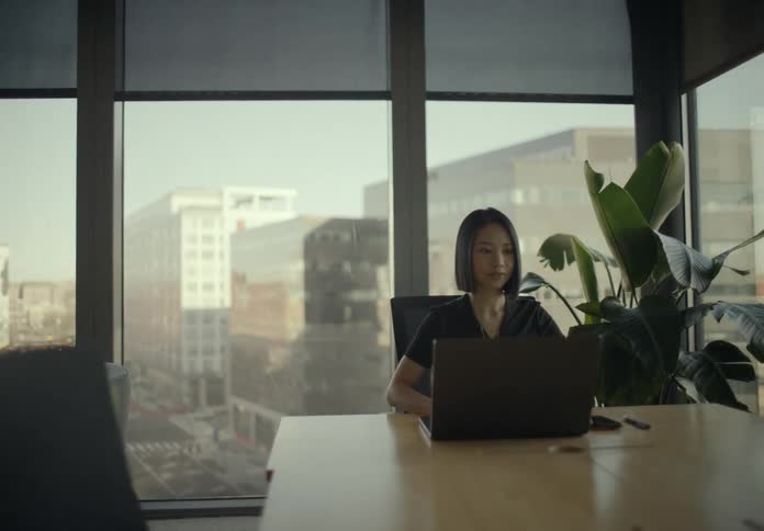 Young adult woman wearing black clothes sitting down in front of a laptop computer over a wooden table in a room with big windows showing a metropolitan area indoors during day time