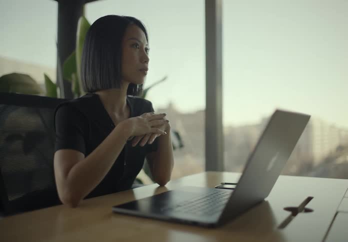 Young businesswoman with short black hair sitting at a wooden desk with a laptop looking thoughtfully out of a large office window during sunset