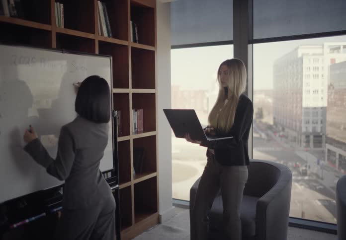 Two professional women collaborate in a modern office with one writing on a whiteboard while the other holds a laptop and provides input the room features a large bookshelf and expansive windows that reveal a clear view of the city streets below