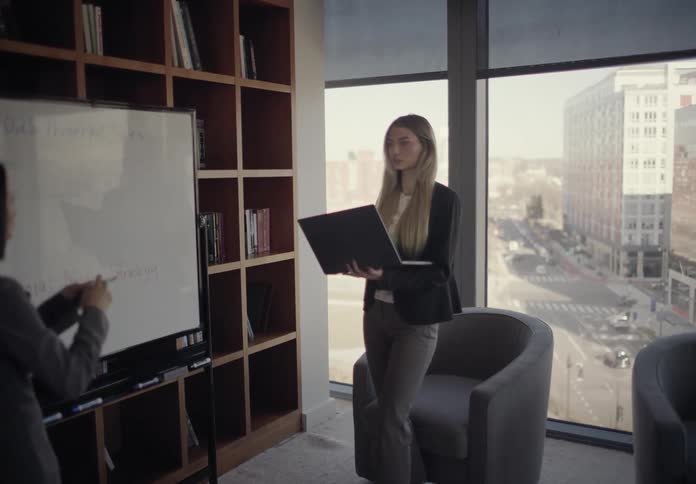 Two professional women collaborate in a modern highrise office and while one speaks near a whiteboard the other one holds a laptop and observes the notes near a large window overlooking the city