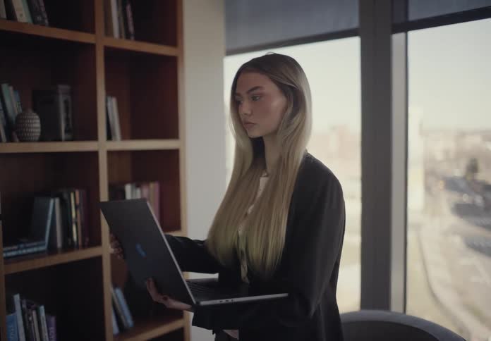 Young adult woman standing up and using a laptop computer held over her arm indoors during day time