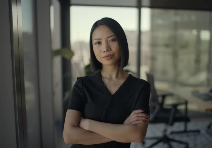 Confident young woman with short black hair standing in a modern office crossing her arms and looking directly into the camera with a neutral expression