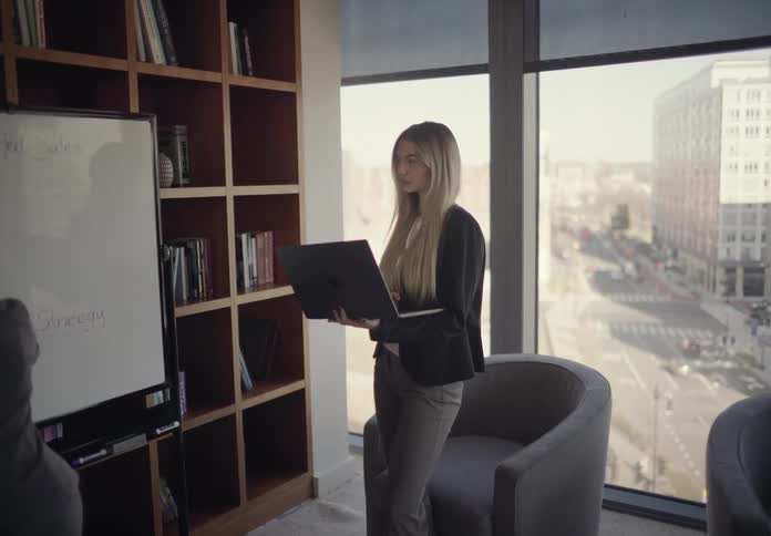 Two professional businesswomen in a bright office one presenting on a whiteboard while the other holds a laptop and looks on with large windows overlooking a city street