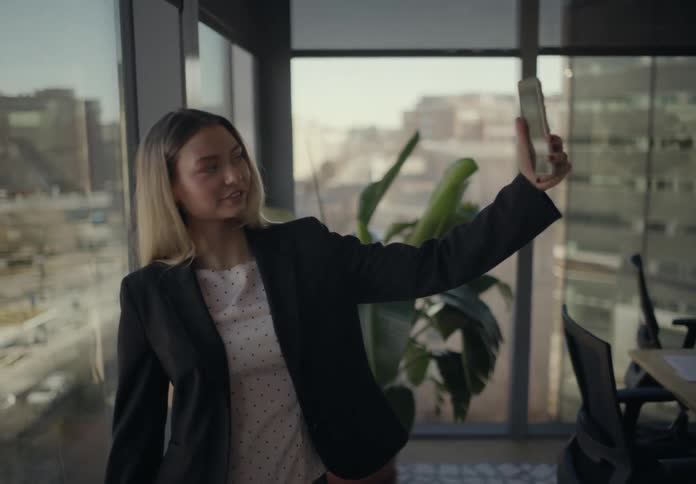 In a sunlit office with large windows overlooking the city a woman in business attire holds up her smartphone at arms length smiling slightly as she appears to take a photo or video near a desk and a tall potted plant