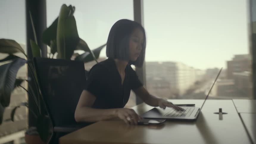Woman sitting at an office table typing on a laptop computer as she stops to look at her phone