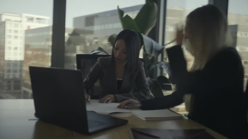 Two businesswomen collaborate at a conference table reviewing and signing documents in a modern office with large windows