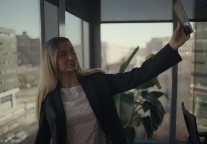 In a modern office with expansive windows and a cityscape beyond a woman in a blazer extends her arm to hold up her smartphone smiling as if capturing a selfie