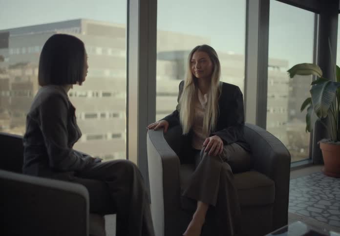 Two young adult women sitting down on armchairs and talking in an office with big windows showing a metropolitan area on the background indoors during day time