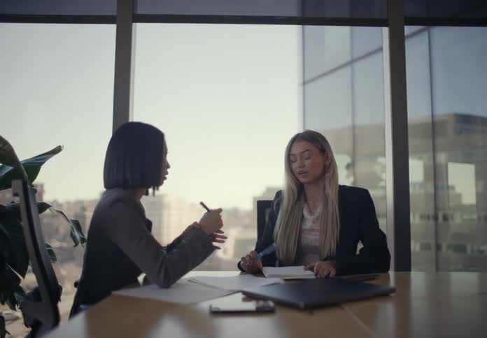 Laptop computer and paperwork on a table as two women sit in a office having a conversation