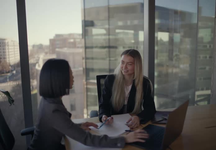 Two women sitting in a conference room at midday in a highrise office building smiling as they discuss together