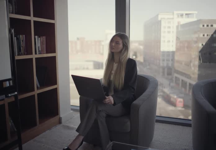 In a sunlit highrise office with a city view a businesswoman writes on a whiteboard while her colleague sits in a grey armchair with a laptop attentively watching and listening to the presentation
