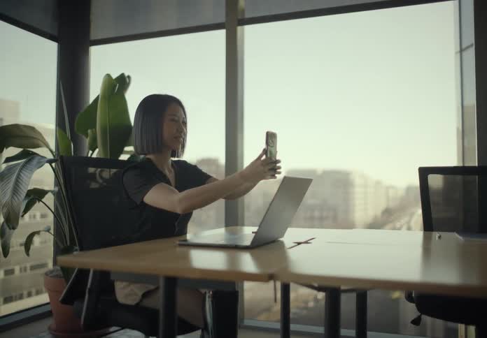 A woman records a video on her smartphone while sitting at her desk in a modern office