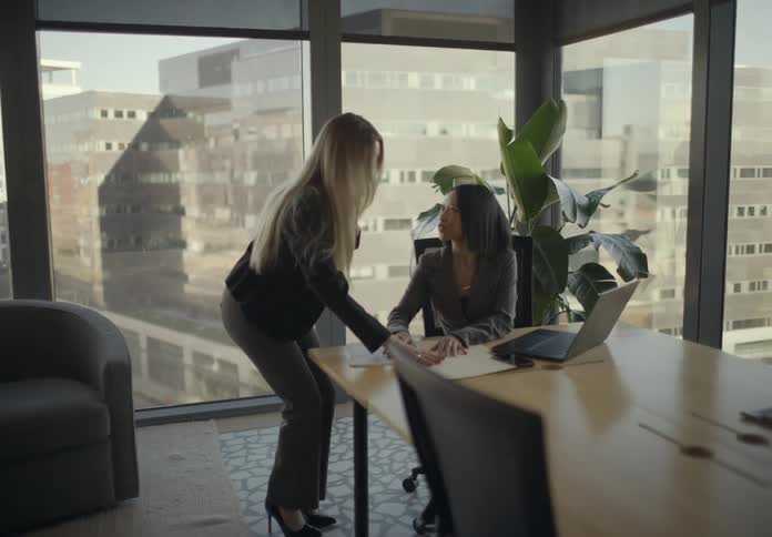 A woman walks across a sunlit modern office to hand a document to a seated colleague who smiles and begins writing on the paper near her laptop while the translucent windows shows an overview of the city