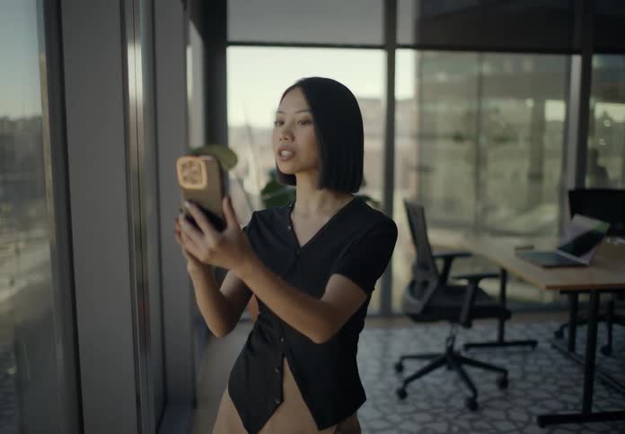 Young businesswoman with short black hair standing in a modern office filming a video on her smartphone while gesturing and speaking toward the camera with sunlit windows in the background