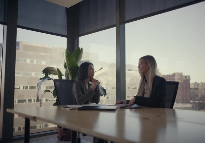 Two young adult women sitting on chair and talking on an office with big windows showing a metropolitan area indoors during day time