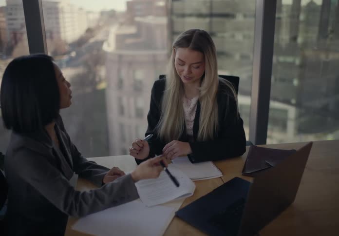 Two professional businesswomen sitting at a desk in a modern office discussing documents and signing paperwork near a large window with a city view