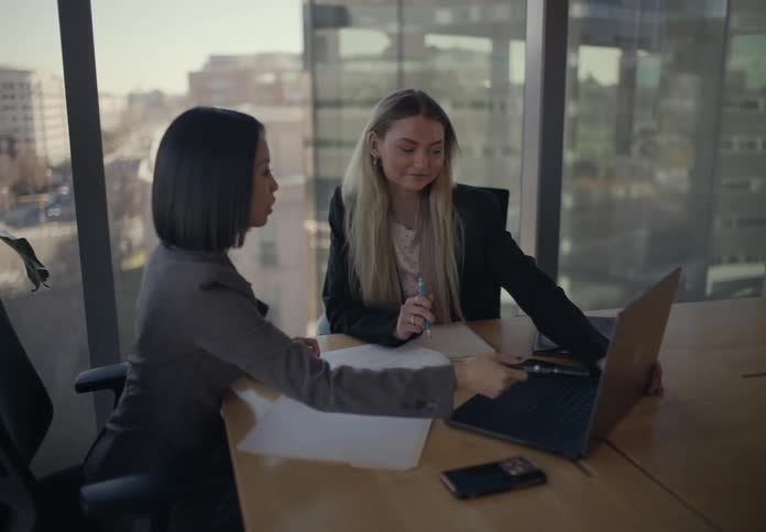Two professional women collaborate in a modern office pointing at a laptop screen while discussing work at a conference table