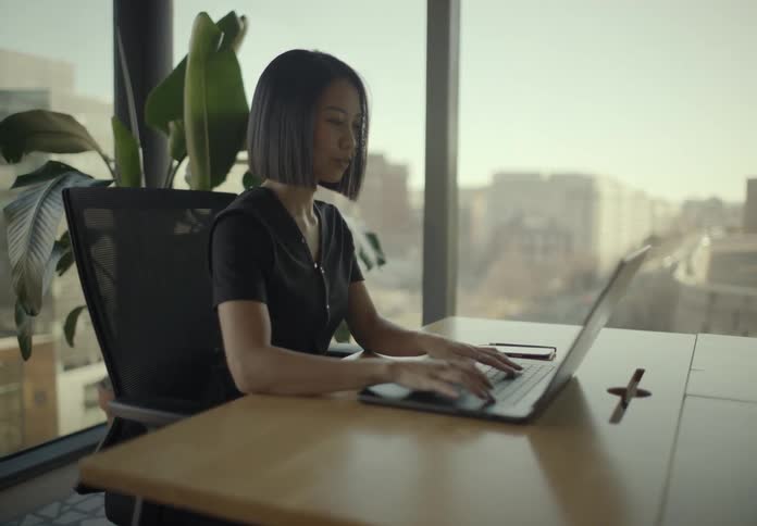 Woman sitting at a table in an office typing on a laptop computer as she stops to look at her phone