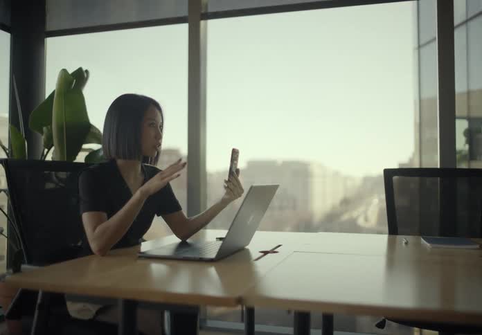In a bright modern conference room with floortoceiling windows a woman sits at a long table using her laptop while holding up her smartphone and gesturing