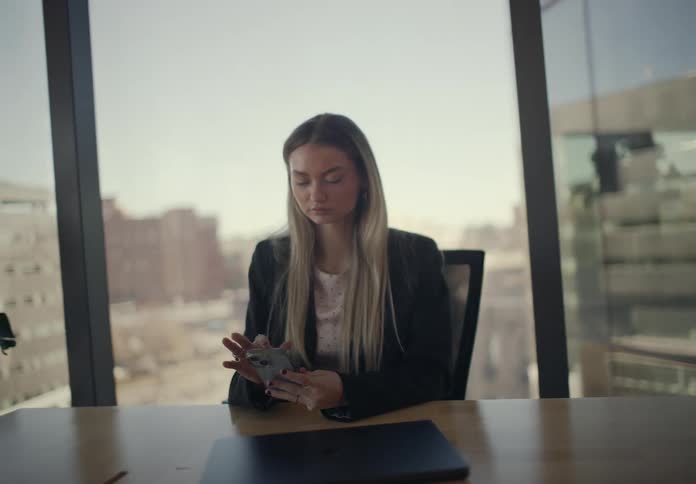 A woman in a black blazer sits at a wooden desk in a highrise office focused on scrolling through her smartphonethe camera slowly zooms out to reveal a large window behind her showing a bright city skyline under clear daylight